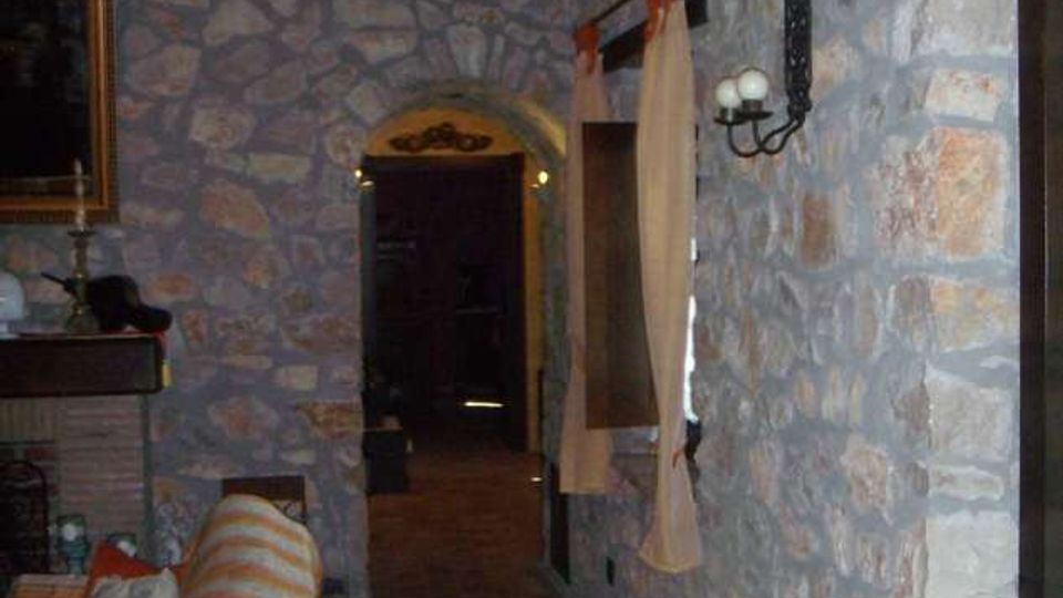 Interno rustico con pareti in pietra a vista e soffitto con travi in legno, pavimento in cotto e corridoio che conduce a una porta ad arco. Sulla destra ci sono una finestra con tende leggere e una applique in ferro battuto; a sinistra si intravede un divano vicino a un camino.