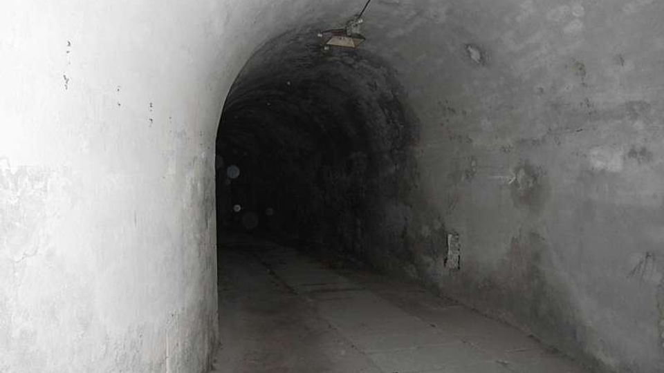 Lungo tunnel sotterraneo in cemento con soffitto a volta e pareti grezze, illuminato debolmente da una lampada a soffitto. Il pavimento è in lastre e il corridoio si restringe verso un’area più buia in fondo.