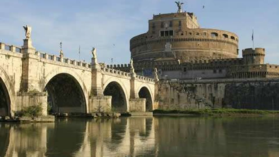 Veduta di Castel Sant’Angelo a Roma con il Ponte Sant’Angelo in primo piano, composto da arcate in pietra e statue lungo il parapetto. Il fiume Tevere scorre sotto il ponte e riflette la fortezza cilindrica con bastioni e mura.