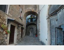 Vicolo in pietra in un borgo storico con una scalinata centrale che sale sotto archi in muratura. Ai lati si vedono facciate in pietra e intonaco, porte in legno e un piccolo balcone con ringhiera e piante.