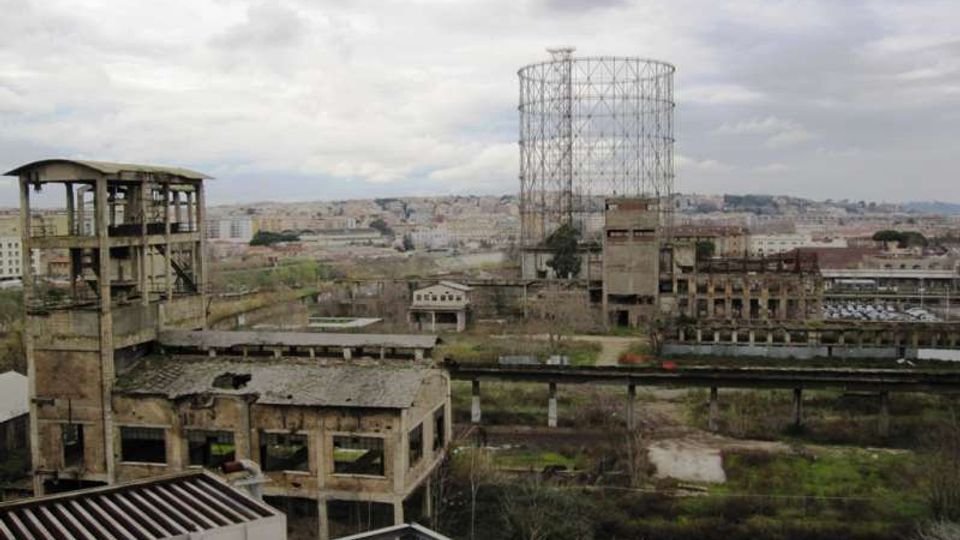 Vista panoramica di un complesso industriale dismesso con capannoni in muratura e strutture metalliche deteriorate, dominato da un grande gasometro cilindrico in traliccio. Sullo sfondo si vede la città sotto un cielo nuvoloso, con aree verdi e binari o passerelle sopraelevate nell’area.