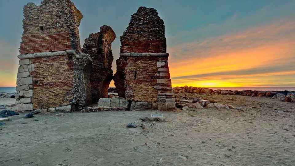 Rovine di una struttura in mattoni e pietra, con due pilastri parzialmente crollati, su una spiaggia sabbiosa vicino al mare. Sullo sfondo si vede la linea dell’orizzonte e un cielo al tramonto con strisce di colore arancione e blu.