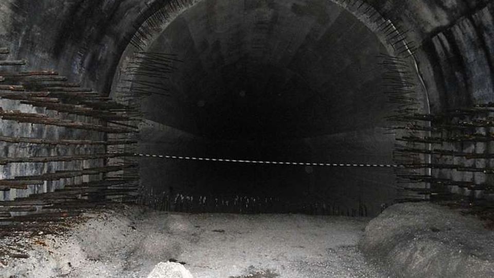 Interno di una galleria o tunnel in cemento con volta semicircolare, pareti annerite e barre metalliche sporgenti sui lati. Il pavimento è coperto di detriti e l’apertura verso il fondo appare buia e profonda.