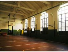 Palestra scolastica interna con campo da basket segnato a terra, soffitto alto con travi a vista e canestro fissato alla parete. Lungo un lato ci sono grandi finestre ad arco con inferriate che illuminano l’ambiente, con pareti bianche e fascia scura nella parte inferiore.