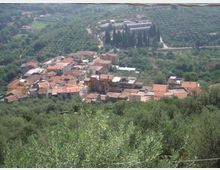 Veduta dall’alto di un piccolo borgo collinare con case addossate e tetti in coppi rossi, circondato da uliveti e vegetazione fitta. Sullo sfondo si vede una strada sinuosa e un complesso di edifici immerso nel verde con alti cipressi.