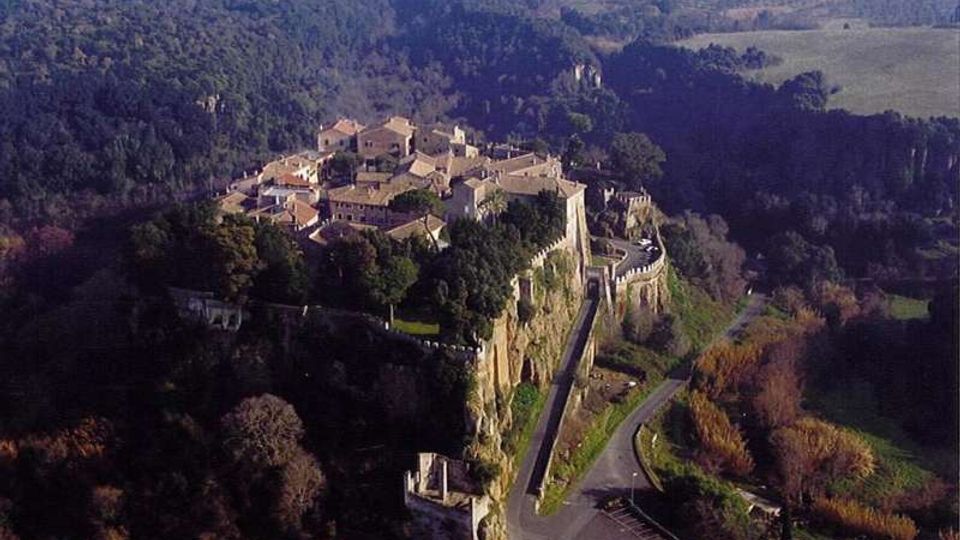 Veduta aerea di un borgo storico arroccato su uno sperone di roccia, con case in pietra dai tetti in coppi e una lunga cinta muraria con bastioni lungo il bordo della scogliera. Una strada curva costeggia la base del promontorio, circondata da boschi e campi agricoli nella campagna circostante.