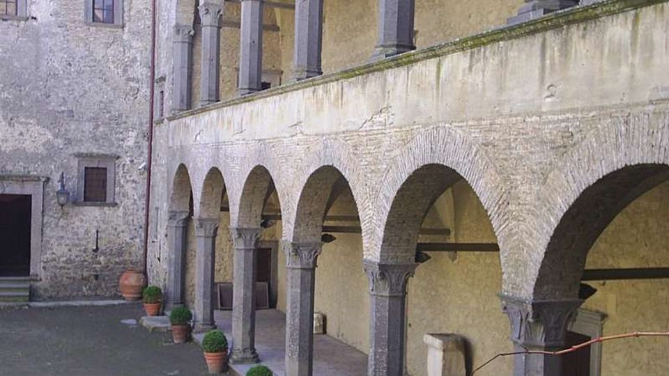Cortile interno di un edificio storico in pietra con portico a due livelli, caratterizzato da file di archi in mattoni sostenuti da colonne grigie. Lungo il porticato sono disposti vasi in terracotta con piante, con muri in pietra e tetti in coppi sullo sfondo.