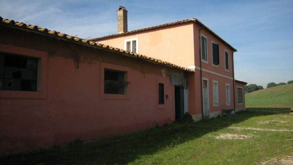 Casale di campagna con facciata rosa e tetto in tegole, composto da un corpo basso con finestre e da un edificio a due piani con persiane e camino. Intorno si vede un prato con sentiero ghiaioso e campi aperti sotto un cielo sereno.
