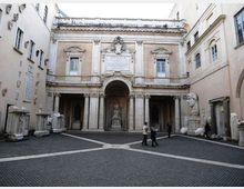 Cortile interno di un complesso storico con pavimentazione in selciato e facciata monumentale in pietra con grande arco centrale e porticato. Ai lati sono disposte statue e frammenti scultorei, tra cui un grande busto, con edifici chiari che chiudono lo spazio su tre lati.