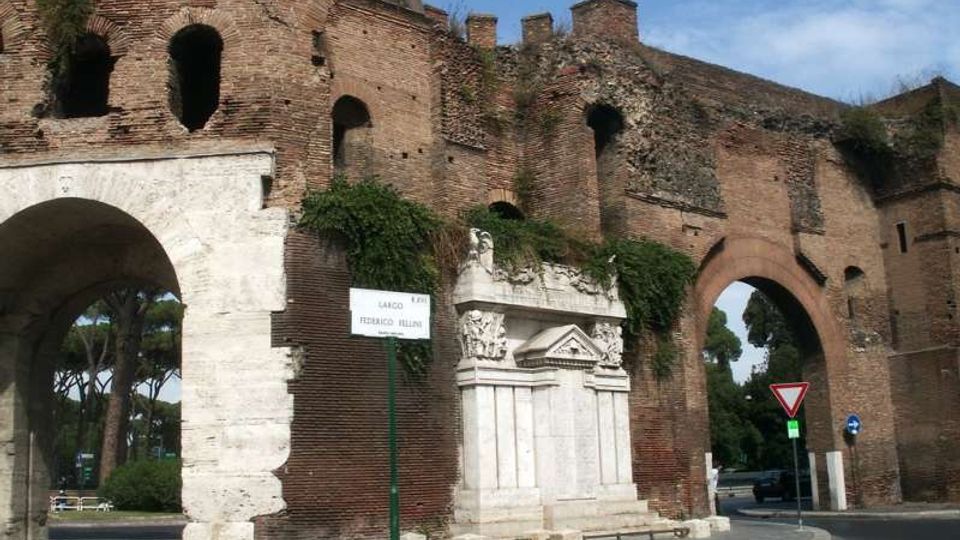Antica porta urbana in mattoni con grandi arcate e muratura parzialmente in rovina, ricoperta in parte da vegetazione. Alla base si vede un monumento in marmo bianco addossato al muro, con strada e segnaletica stradale sullo sfondo.