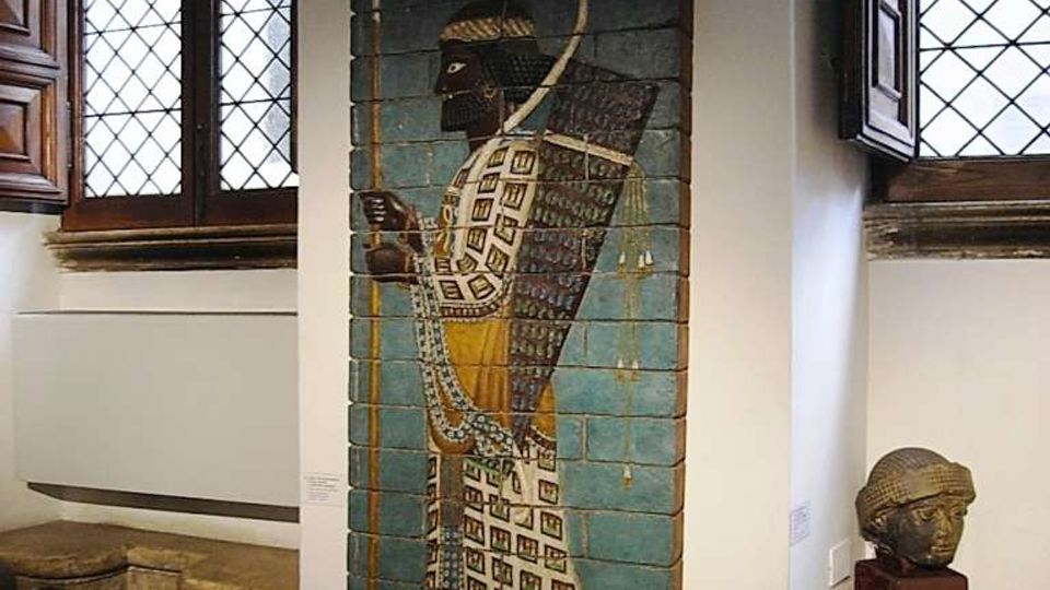 Interno di museo o galleria con pareti bianche e grandi finestre ad arco con vetri a losanghe e scuri in legno. Al centro è esposto un pannello verticale decorato in stile antico su supporto azzurro, con una piccola scultura su basamento accanto e gradini in pietra sul lato.