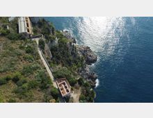 Veduta aerea di un promontorio roccioso sul mare con vegetazione mediterranea, una strada stretta e alcuni edifici in muratura lungo la costa. Il litorale presenta scogliere a picco e acque blu con riflessi di luce.