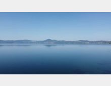 Ampia veduta di un lago dalle acque calme sotto un cielo limpido, con una catena di colline e una costa lontana all’orizzonte.