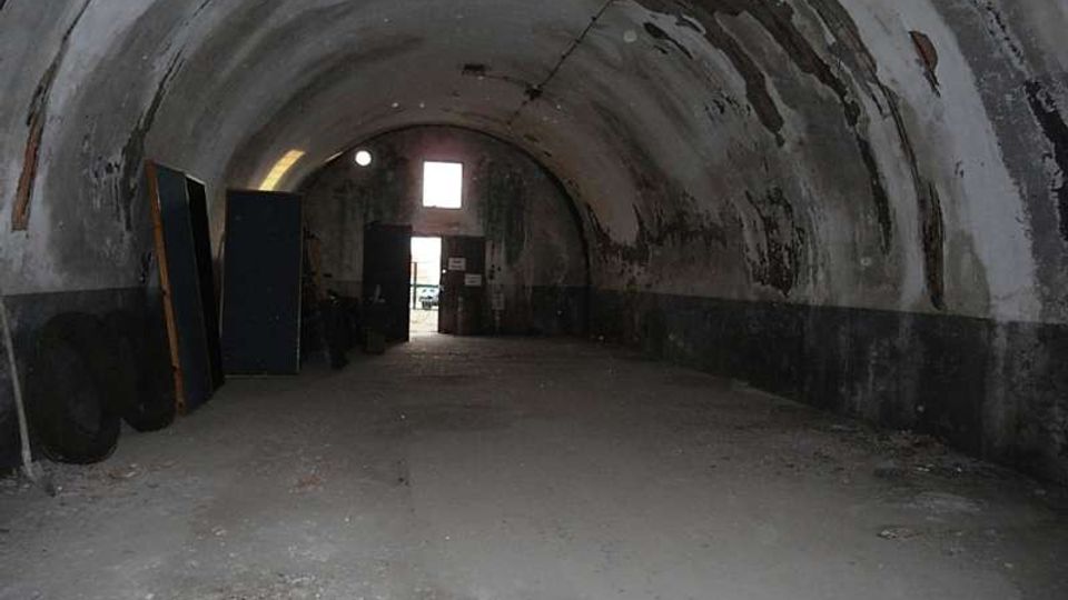 Interno di un grande tunnel o rifugio sotterraneo con volta a botte in cemento, pareti scrostate e pavimento polveroso con detriti. In fondo si vede un’apertura rettangolare verso l’esterno e alcune grandi porte o pannelli appoggiati lungo il lato sinistro.