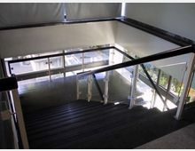 Interno di un vano scala moderno con gradini scuri e parapetti in vetro e metallo, affacciato su grandi finestre che lasciano entrare molta luce naturale. Le vetrate permettono la vista dell’esterno e di un’area con vegetazione.