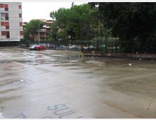 Ampio piazzale asfaltato bagnato dalla pioggia con segni e scritte sul pavimento, delimitato da una recinzione metallica. Sullo sfondo si vedono auto parcheggiate, alberi alti e palazzi residenziali.