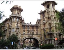 Palazzo storico in stile eclettico con facciate riccamente decorate, due torri angolari e un grande arco centrale che attraversa la strada. In primo piano si vedono alberi, auto parcheggiate e segnaletica stradale in un contesto urbano.