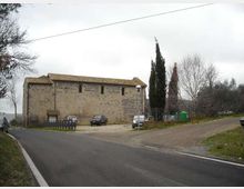 Edificio rurale in pietra con tetto a falde in tegole, affacciato su una strada asfaltata che curva e si biforca. Davanti si vede un piccolo parcheggio sterrato con recinzione, lampioni e alberi alti e spogli sullo sfondo.
