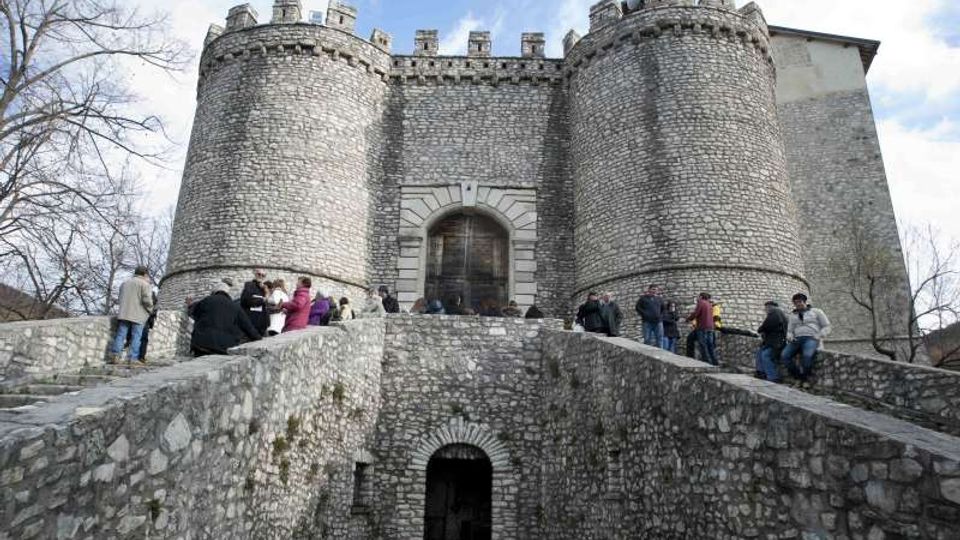 Castello in pietra con due grandi torri cilindriche merlate e portale centrale, visto dal ponte in muratura che conduce all’ingresso. Intorno si notano mura in pietra, un fossato sottostante e alberi spogli sullo sfondo sotto un cielo parzialmente nuvoloso.