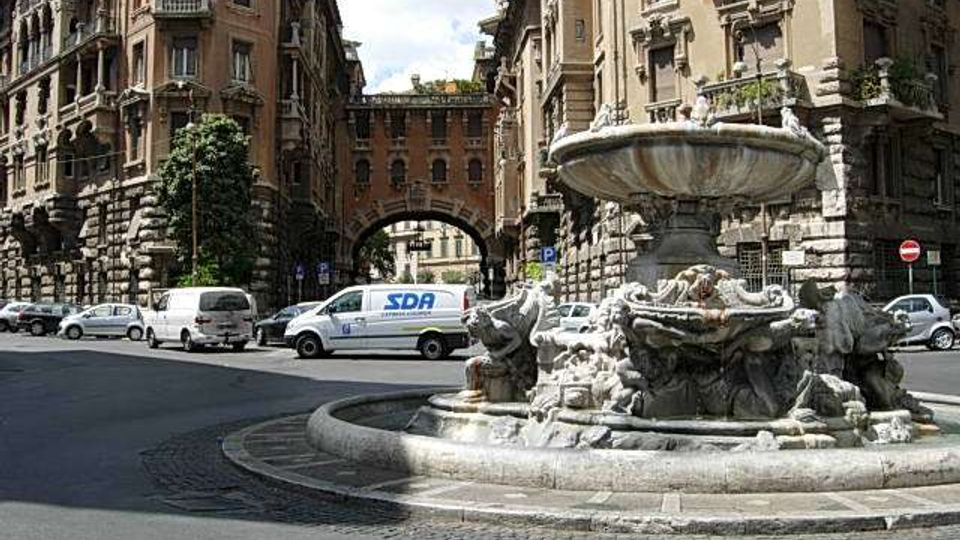 Rotonda urbana con grande fontana in pietra scolpita al centro, circondata da strada asfaltata. Sullo sfondo si vedono palazzi storici in stile monumentale con facciate in muratura, balconi e un arco che collega due edifici, con auto parcheggiate lungo i lati.