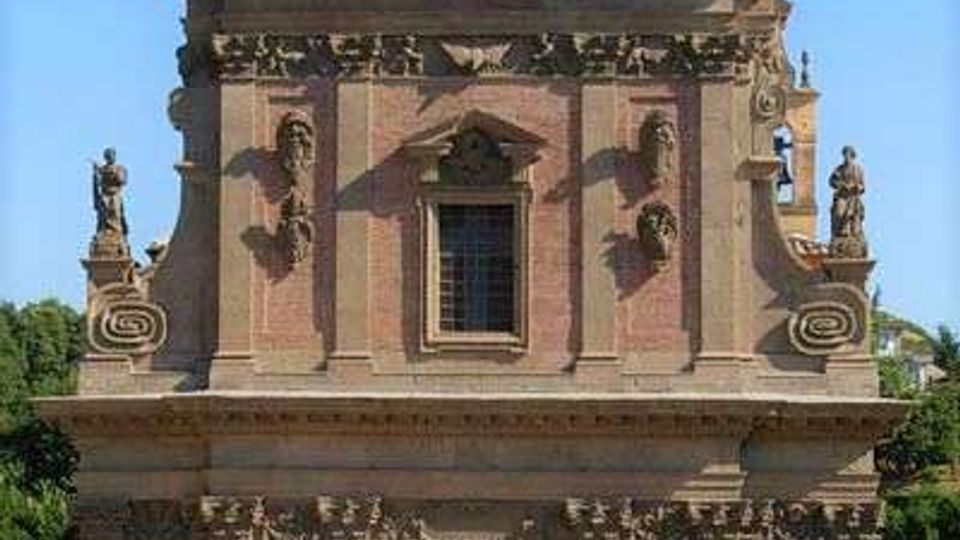 Facciata frontale di una chiesa in mattoni con elementi decorativi in pietra, portale centrale e finestra sopra l’ingresso. Il prospetto è sormontato da un timpano con statue e pinnacoli, su sfondo di cielo sereno e alberi ai lati.