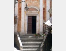 Ingresso di un edificio storico con ampia scalinata in pietra che conduce a un portale con cornice in marmo e porta in legno scura parzialmente aperta. La facciata intonacata presenta nicchie verticali, lampioni a muro e alcune luci decorative, con impalcatura e cartelli sul lato destro.