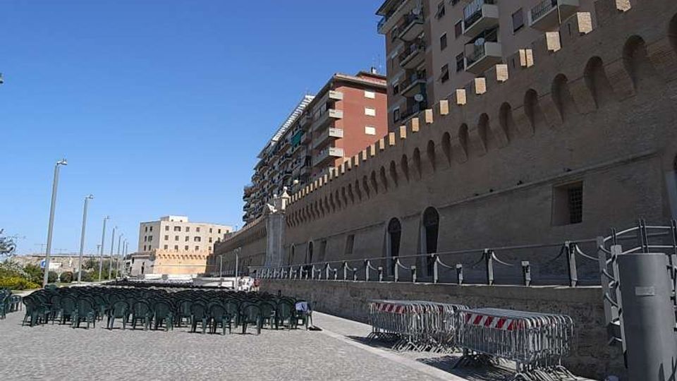 Ampia area pavimentata in pietra lungo una lunga muraglia in mattoni con arcate e merlature, affiancata da edifici residenziali alti con balconi. In primo piano si vedono file di sedie impilate, transenne metalliche e lampioni, sotto un cielo sereno.