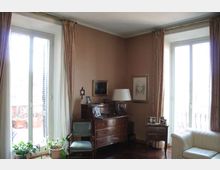 Interno di un soggiorno con due grandi portefinestre affacciate su un balcone, tende lunghe e pareti color tortora. Al centro ci sono un mobile in legno tipo secretaire con lampada e quadri appesi, una sedia, un piccolo tavolino con cornici e una poltrona chiara; alcune piante sono vicino alla finestra.