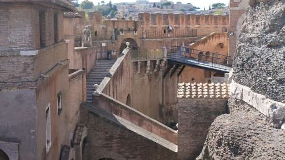 Vista di un complesso fortificato in mattoni con mura merlate, archi e passaggi sopraelevati. Un ponte pedonale metallico collega le strutture, con tetti in tegole e pareti in pietra visibili ai lati.