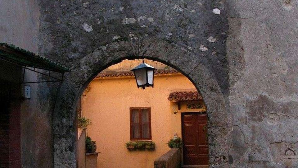 Arco in pietra di un vicolo medievale tra edifici intonacati, con una lanterna appesa al centro. Oltre l’arco si vede un cortile con scala in cemento che porta a una porta in legno e una finestra su una facciata color ocra.