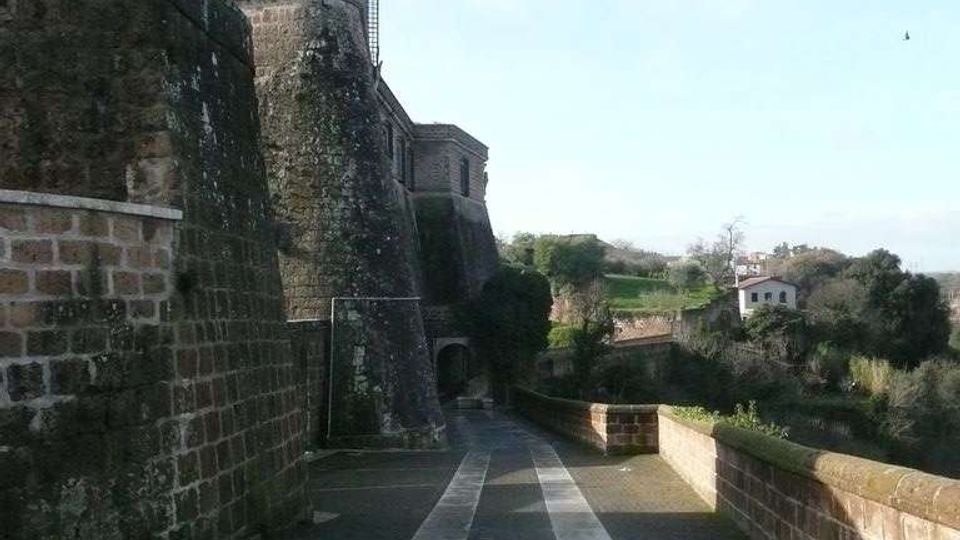 Camminamento in pavé lungo le mura di una fortificazione in pietra, con torre curva e pareti alte ricoperte in parte di vegetazione. Il percorso è affiancato da un parapetto in muratura e si apre verso un panorama di alberi e case sullo sfondo.