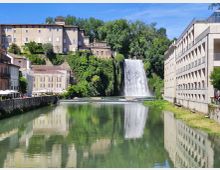 Veduta urbana lungo un fiume calmo con una grande cascata che scende da una scarpata verde. Ai lati si vedono edifici storici su una collina e un lungo edificio moderno sul lungofiume, con riflessi nell’acqua.