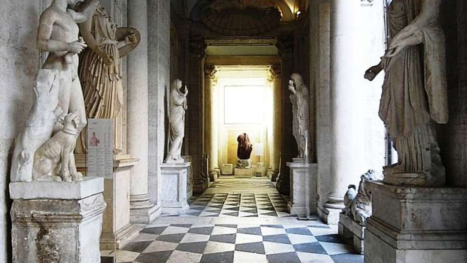 Corridoio monumentale in pietra con archi e colonne, pavimento a scacchi bianco e nero e statue classiche su piedistalli lungo entrambi i lati. In fondo si apre un vano luminoso con una statua centrale, illuminato da luce naturale.