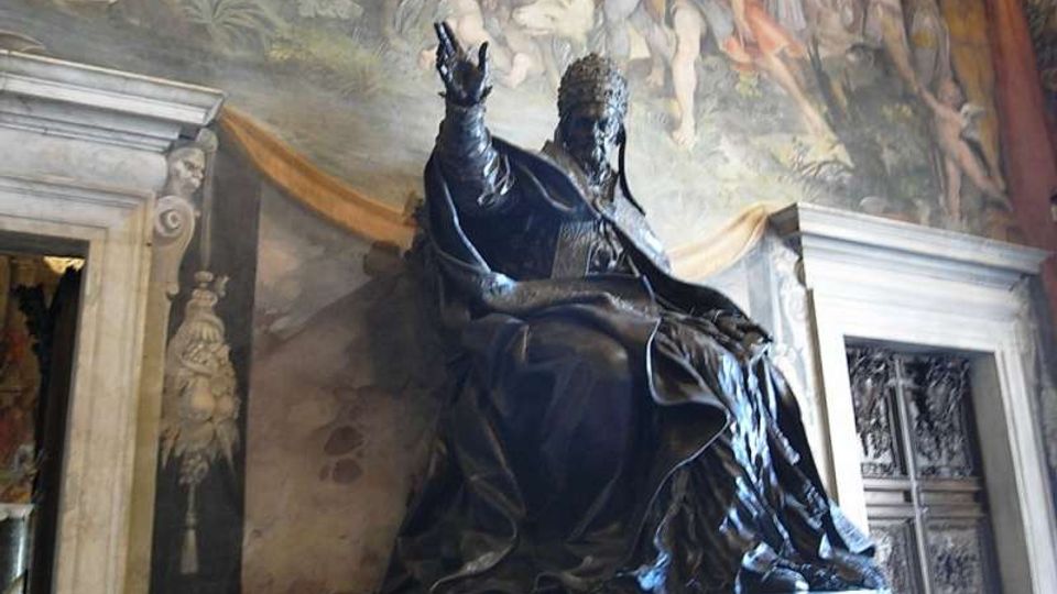 Statua monumentale in bronzo di una figura seduta su un alto basamento, collocata in un interno storico con grandi affreschi murali sullo sfondo. Ai lati si vedono portali in pietra con cornici decorative e una porta a pannelli intagliati.