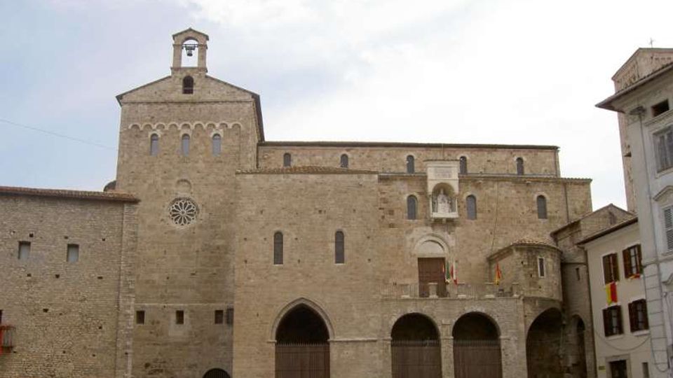 Facciata in pietra di una chiesa medievale con campanile a vela, rosone e finestrine ad arco; alla base si vedono portici con archi e grandi ingressi. L’edificio si affaccia su una piazza, con altri palazzi ai lati.