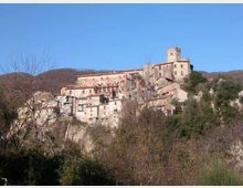 Borgo storico arroccato su un pendio, con case in pietra addossate tra loro e una torre quadrata in cima al complesso. Intorno si vedono colline boscate e vegetazione in primo piano sotto un cielo limpido.