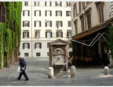 Piccola piazza acciottolata tra edifici storici con persiane e facciate chiare, con una fontana in pietra al centro. A sinistra una parete ricoperta di edera e a destra un portico con tende davanti a un locale.
