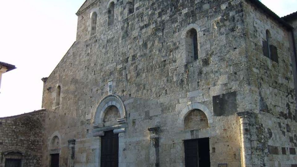 Facciata di una chiesa romanica in pietra con grandi muri spessi, archi a tutto sesto e portali scuri incassati. La struttura presenta piccole finestre ad arco nella parte alta e un tetto a due falde, affacciata su una piazza lastricata.