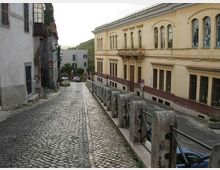 Strada in discesa con pavimentazione in sampietrini, affiancata da edifici storici e da una balaustra in pietra con corrimano metallico. Sulla destra si vede un grande edificio color crema con finestre ad arco, con alcune auto parcheggiate lungo la via.
