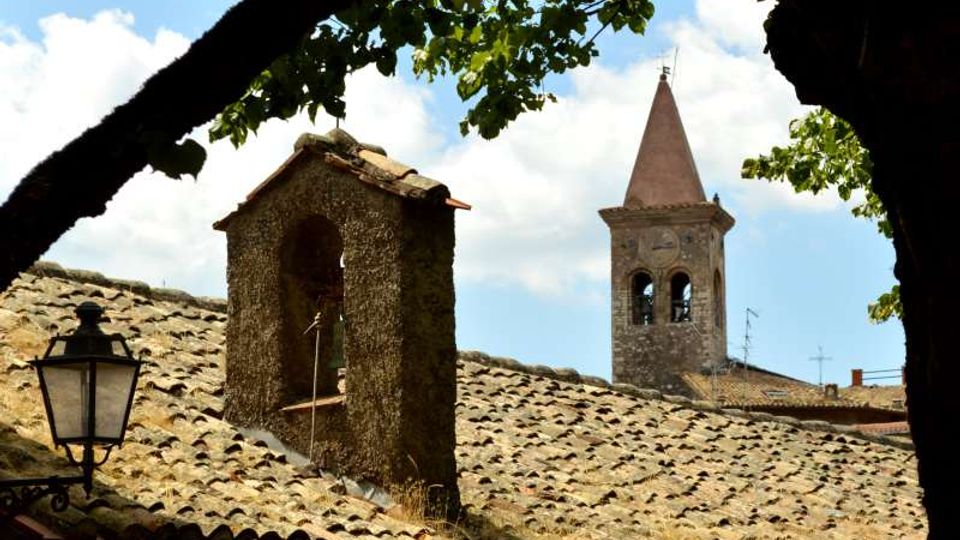 Tetti in coppi di un borgo storico con un piccolo campanile in pietra e, sullo sfondo, una torre campanaria con cuspide. L’inquadratura è incorniciata da rami e foglie di albero, con un lampione in ferro in primo piano.