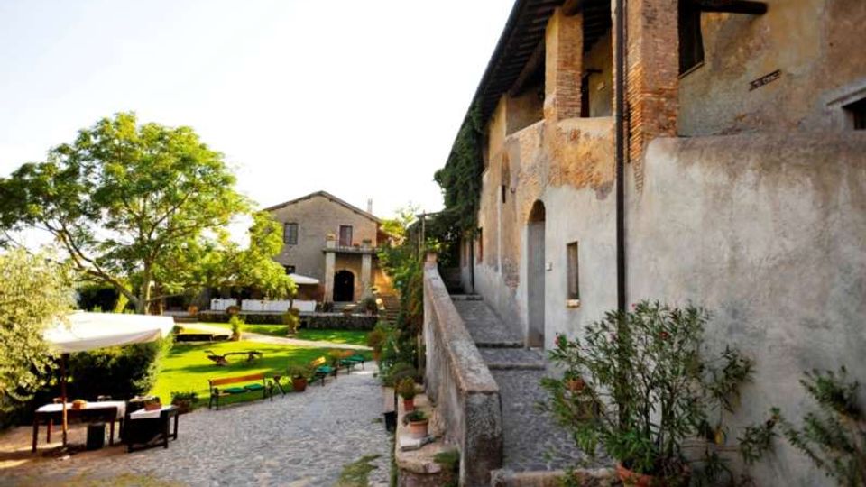 Cortile di un casale rustico in pietra e intonaco, con lungo portico ad archi e camminamento rialzato lungo la facciata. In primo piano un vialetto in ciottoli conduce a un’area verde con alberi, tavoli e panche all’aperto, e un edificio secondario sullo sfondo.