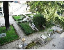 Giardino formale con vialetti in ghiaia e aiuole geometriche bordate da siepi basse, con una piccola fontana o vasca circolare al centro e sculture in pietra. Alberi alti e un edificio chiaro con scala esterna fanno da sfondo.