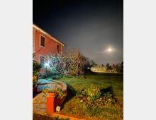 Giardino notturno accanto a una casa in muratura rossa, con vialetto in pietra, aiuole e piccoli lampioni. Sul prato si vede una pergola coperta da rami spogli e, sullo sfondo, alberi e recinzione sotto un cielo con la luna.