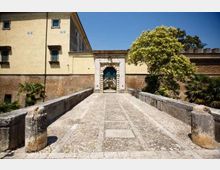 Ponte in pietra con pavimentazione a mosaico che conduce a un portale ad arco in muratura, inserito nelle mura di un edificio storico. Ai lati si vedono muri in mattoni e alberi, con cielo sereno sullo sfondo.