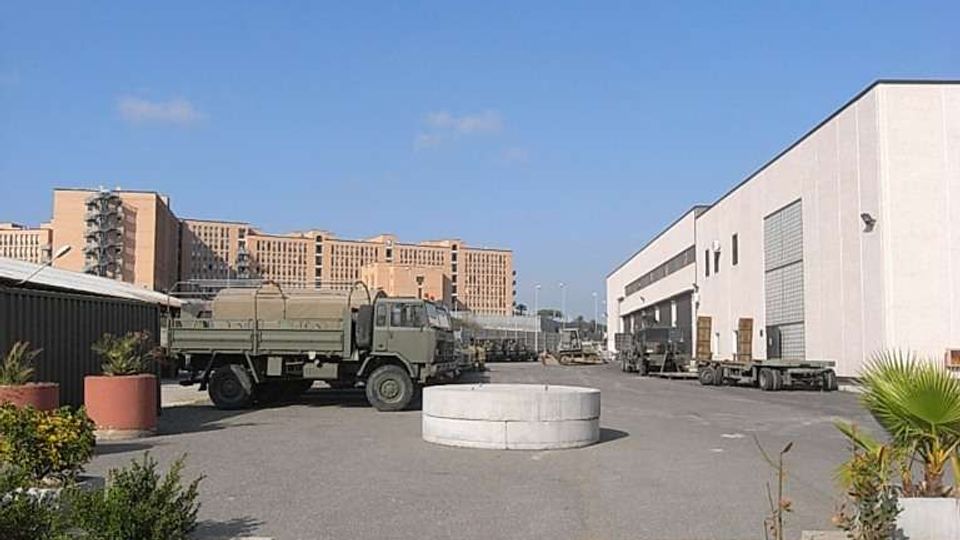 Ampio piazzale asfaltato di un’area industriale, con diversi camion parcheggiati e un grande capannone bianco con portoni sul lato destro. In primo piano ci sono aiuole con piante e una struttura circolare bassa al centro del piazzale, con edifici più alti sullo sfondo sotto un cielo sereno.