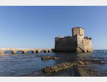 Fortezza costiera in pietra su un isolotto, con torre quadrata e mura merlate, collegata alla riva da un ponte basso ad archi. In primo piano si vede il mare calmo con scogli affioranti e un ampio cielo sereno sullo sfondo.