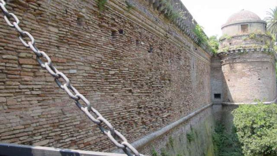 Mura di una fortificazione in mattoni viste da un ponte con parapetto e una grossa catena metallica in primo piano. Sullo sfondo si nota una torre cilindrica con cupola e vegetazione lungo il fossato.