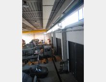 Interno di un grande capannone industriale tipo officina, con soffitto alto in metallo e lucernari laterali che illuminano l’ambiente. Lungo le pareti ci sono ampie porte scorrevoli e all’interno sono presenti vari veicoli e macchinari pesanti parcheggiati sul pavimento in cemento.