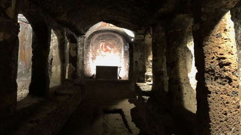 Interno di una cripta o cappella sotterranea scavata nella roccia, con navata centrale e pilastri grezzi che sostengono una volta bassa. In fondo si vede un piccolo altare davanti a un’abside con affresco, illuminata da una luce proveniente dall’alto.