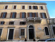 Facciata di un edificio storico a più piani con intonaco chiaro, finestre rettangolari con scuri in legno e un piccolo balcone in pietra sopra un portale ad arco con cornice bugnata. Al piano strada si vedono grandi porte scure e finestre con inferriate, con cielo azzurro sullo sfondo.